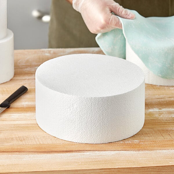 A gloved hand cuts a white round cake dummy with a knife.