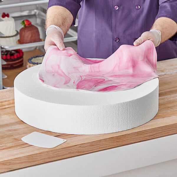 A person using a pink and white fabric to decorate a large round white cake with a Baker's Lane foam cake dummy.