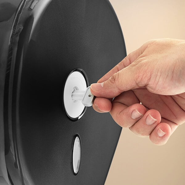 A hand using a Lavex Plastic Key to open a black toilet paper dispenser.