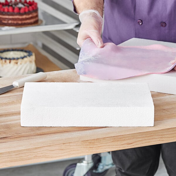 A person cutting a Baker's Lane rectangular foam cake dummy with a knife.