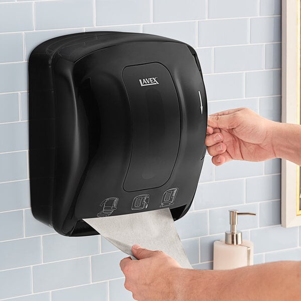 A person using a Lavex Black Lever-Activated Paper Towel Dispenser on a tile wall.