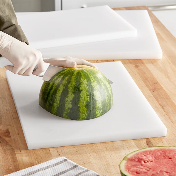 A person using a Choice white polyethylene cutting board to cut a watermelon.