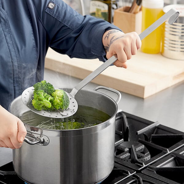 A person using a Choice stainless steel skimmer to cook broccoli in a pot.