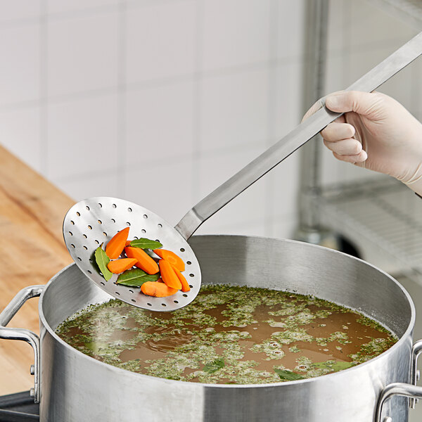 A hand holding a long metal Choice 6" Two-Piece Stainless Steel Skimmer stirring vegetables in a pot.