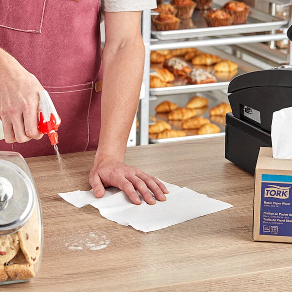 A person using a Tork white light-duty paper wiper to clean a table.