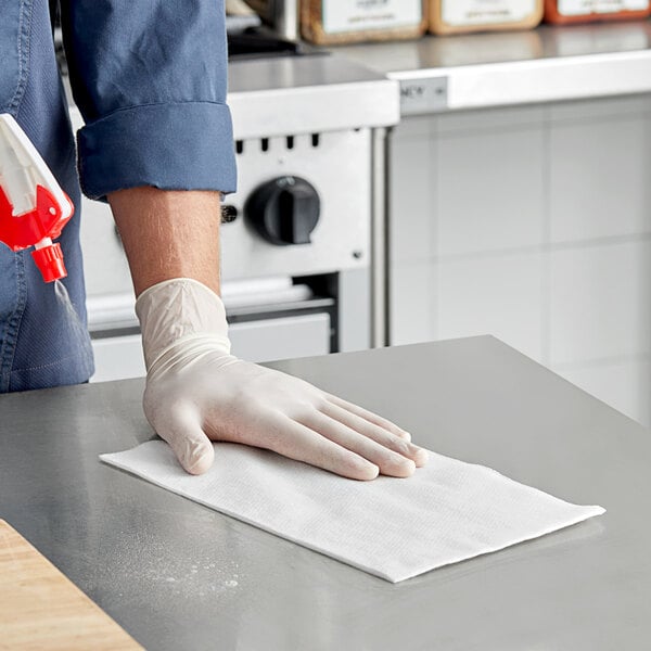 A person wearing a glove and using a Tork white food service towel to clean a surface.