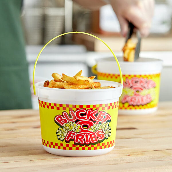 A person holding a 48 oz. plastic French fry bucket on a table of fries.