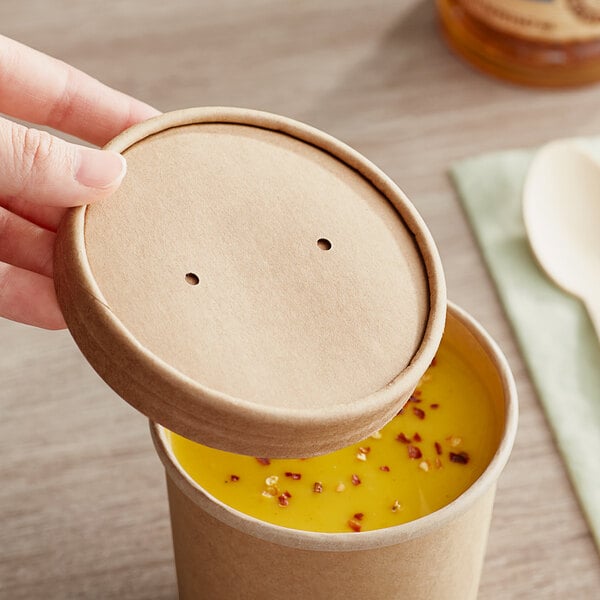 A hand holding a round brown Kraft paper container of soup with a lid.