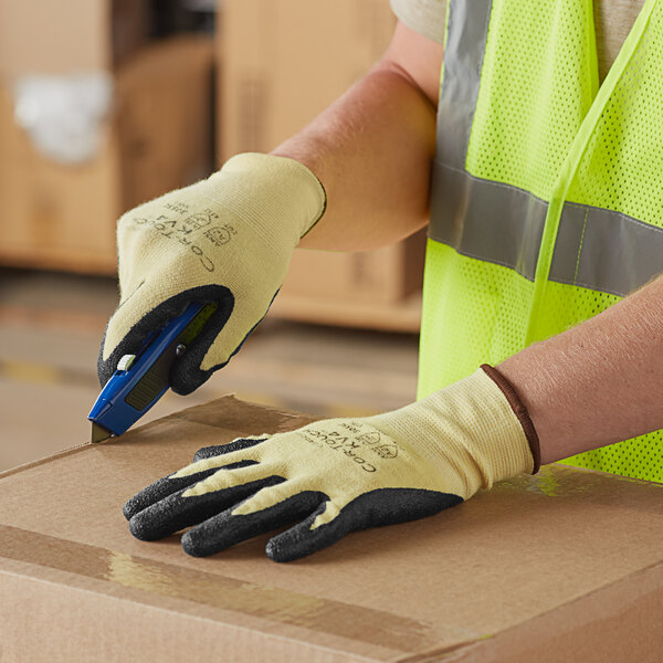 A person wearing Cordova cut-resistant gloves and a safety vest uses a blue and black cutter to cut a cardboard box.