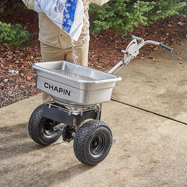 A person pours salt from a bag into a Chapin stainless steel salt spreader.