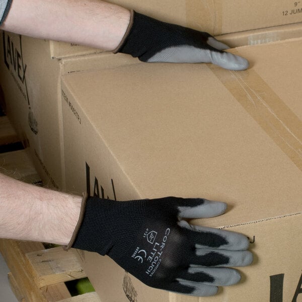 A person wearing Cordova black nylon gloves with gray polyurethane palm coating holding a box.