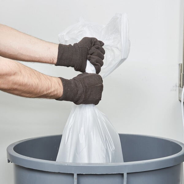 A person in Cordova standard brown jersey gloves holding a plastic bag in a trash can.