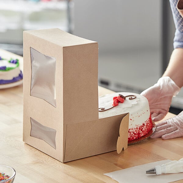 A woman using a white gloved hand to cut a cake in a Baker's Mark Kraft cake box with a window.