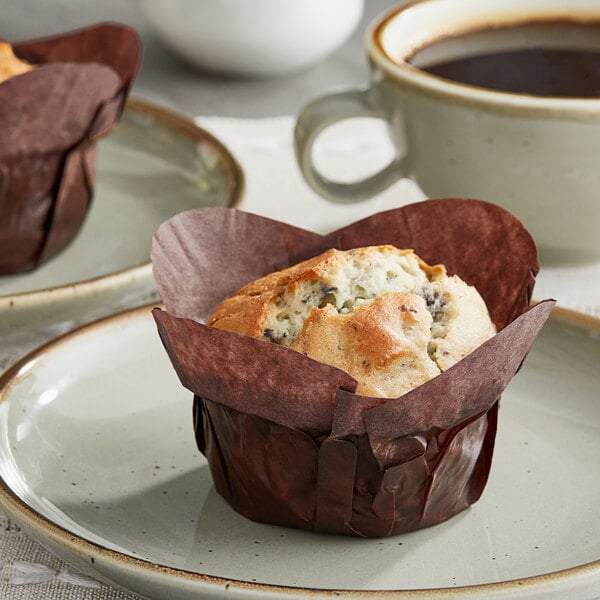 Two Baker's Mark chocolate brown muffins in wrappers on a white plate with a cup of coffee.