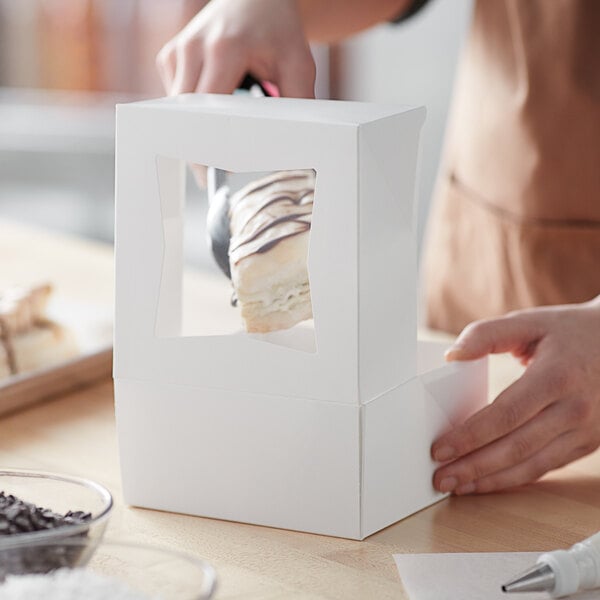 A person holding a piece of cake in a white Baker's Mark bakery box with a window.