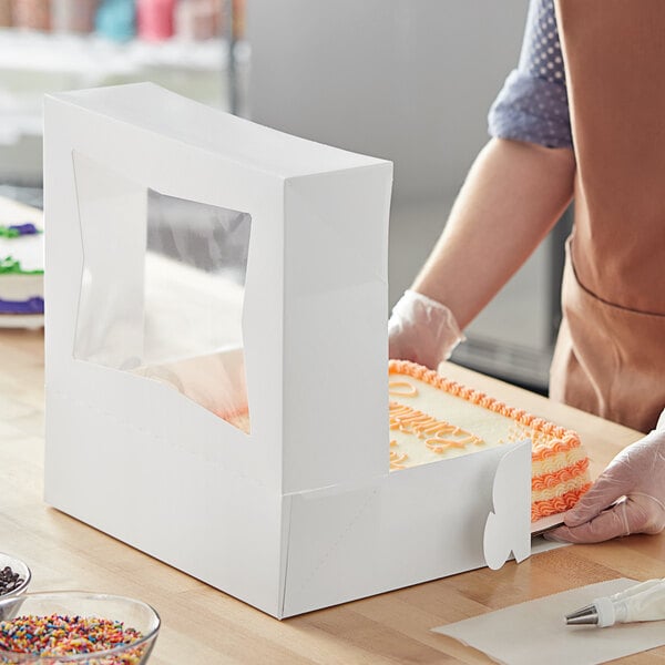 A person in an apron and gloves using a knife to cut a cake in a white Baker's Mark box.