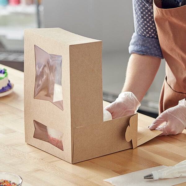 A person wearing gloves and a blue shirt opens a Baker's Mark Kraft bakery box with a window to reveal a cake inside.