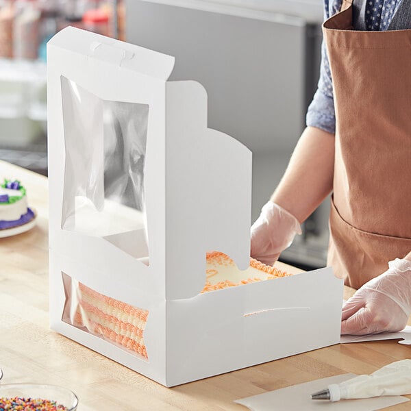 A person wearing gloves and a brown apron cutting a cake in a white Baker's Mark box with orange frosting on top.