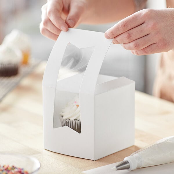 A person opening a Baker's Mark white window cupcake box with a cupcake inside.