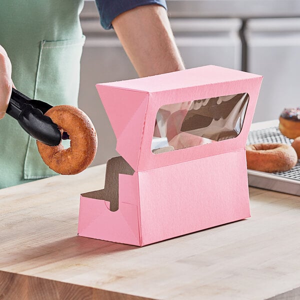 A person using black tongs to place a donut in a pink Baker's Mark bakery box with a window.