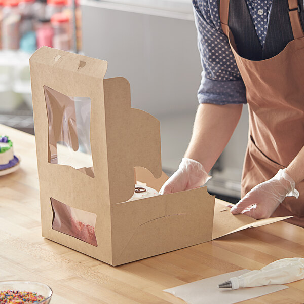 A woman in an apron cutting a cake in a Baker's Mark bakery box on a counter.