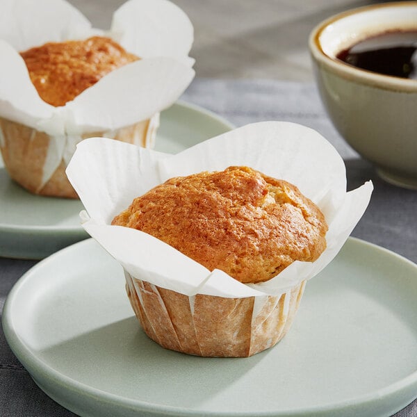 Two Baker's Mark white paper-wrapped muffins on plates with a cup of coffee.