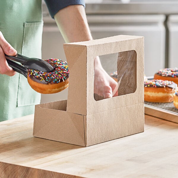 A person holding a chocolate covered doughnut with sprinkles in a Kraft window bakery box.