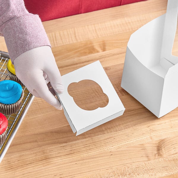 A person in pink gloves holding a Baker's Mark cupcake box with a cut-out.