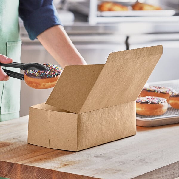 A person using tongs to pick up a donut with sprinkles from a Baker's Mark bakery box.