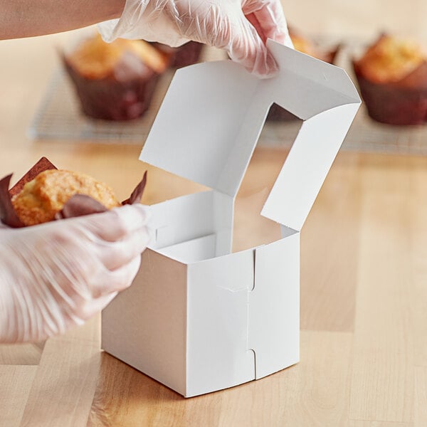 A gloved hand places a muffin in a white window box with a plastic lid.