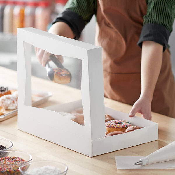 A person wearing a brown apron putting a donut into a white Baker's Mark bakery box.