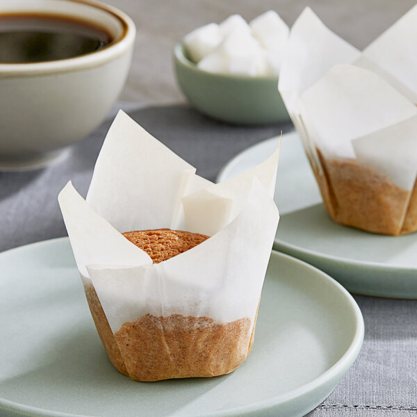 Two Baker's Mark white tulip baking cups with muffins on plates next to a cup of coffee.