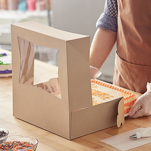 A person in a brown apron and gloves opening a Baker's Mark Kraft quarter sheet cake box with a window.