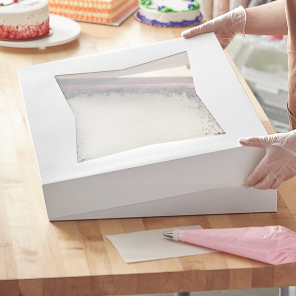 A person holding a Baker's Mark white box with a clear window over a cake.