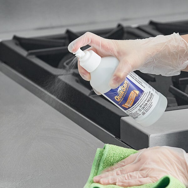 A hand in a white glove spraying Noble Chemical QuikSan surface sanitizer on a kitchen counter.