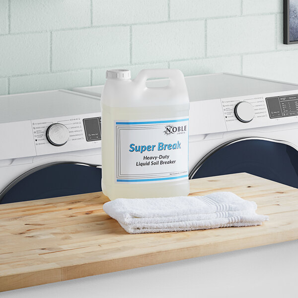 A white jug of Noble Chemical Super Break Alkaline Laundry Soil Breaker on a wood surface next to a washing machine.