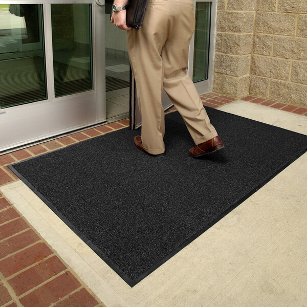 A person is stepping onto a large, dark-colored floor mat placed outside a glass door entrance.