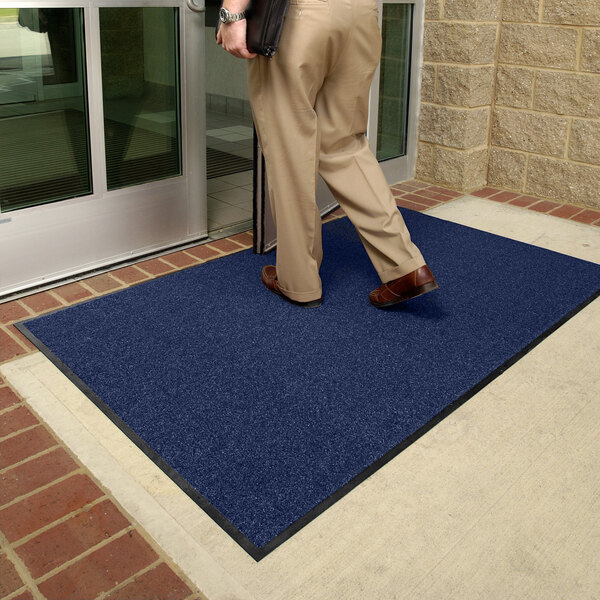 A person is stepping onto a large, rectangular navy blue mat with a black border placed outside a glass door entrance.
