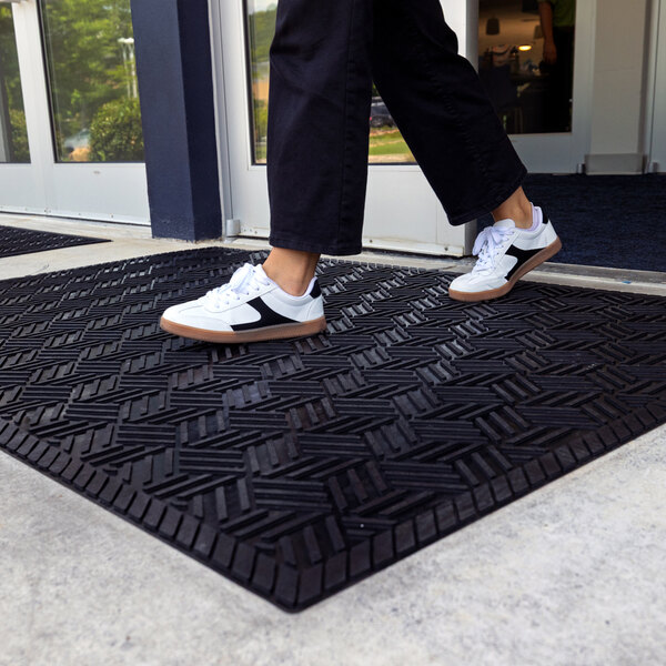 A person wearing white sneakers and black pants is stepping onto a large black rubber mat with a geometric pattern, placed outside a building entrance.