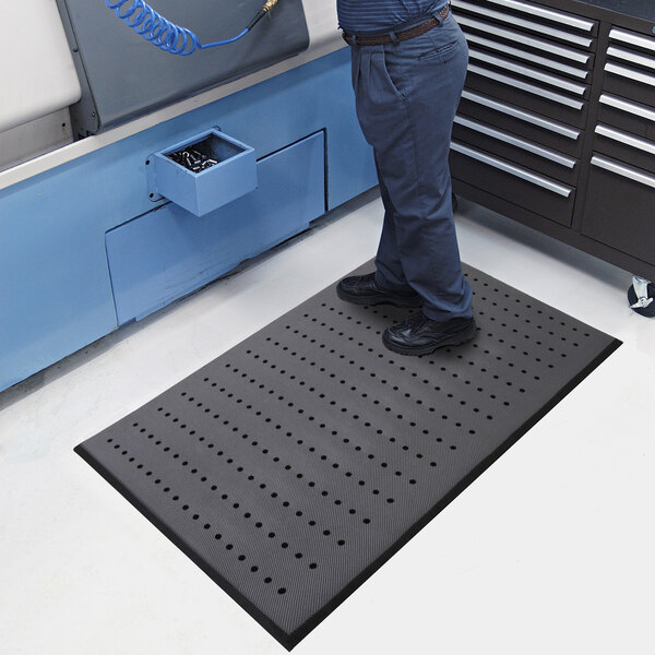A person is standing on a black mat with evenly spaced drainage holes and beveled edges, placed on a light-colored floor in an industrial or workshop setting.