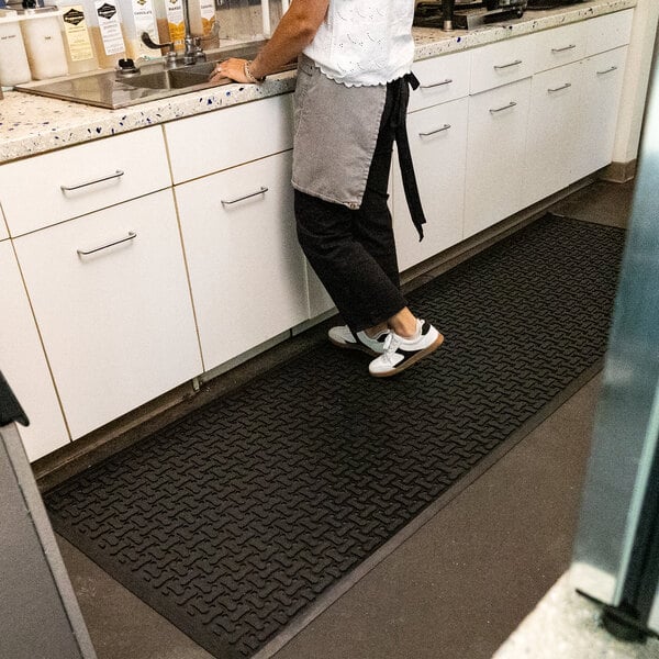 A person wearing an apron stands on a large black rubber mat with a textured pattern and beveled edges, placed in front of a kitchen counter.