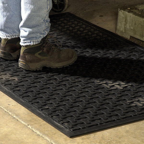 A person wearing work boots and jeans is standing on a black textured rubber mat with a beveled edge on a concrete floor.