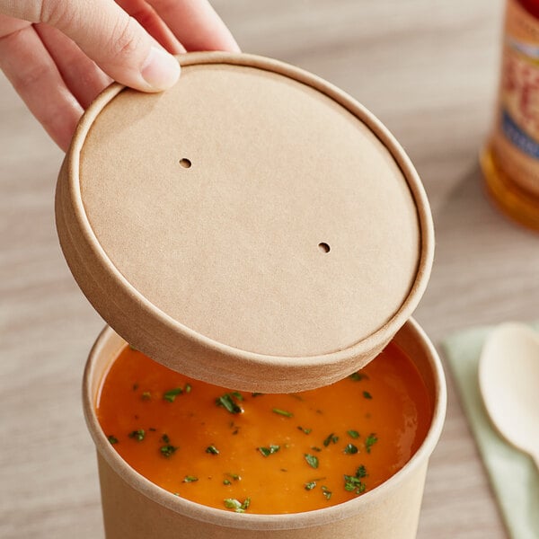 A hand holding a Choice 32 oz. Kraft Paper Soup Container with soup and a vented lid.