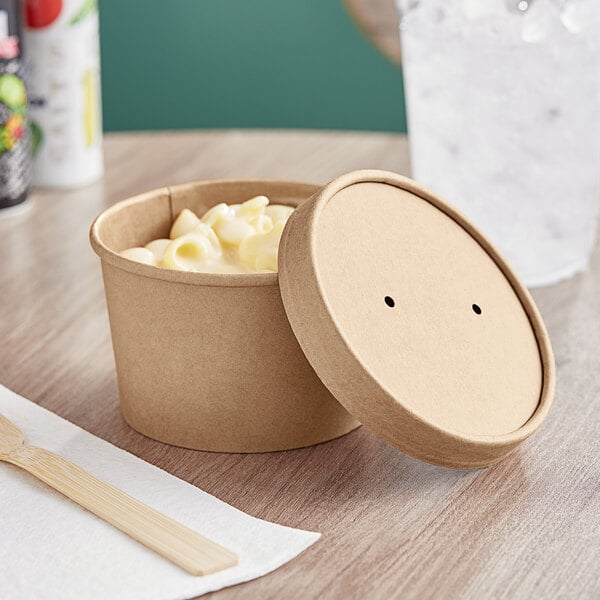 A round Kraft paper container with a vented paper lid on a table with a container of macaroni and cheese and a wooden spoon.