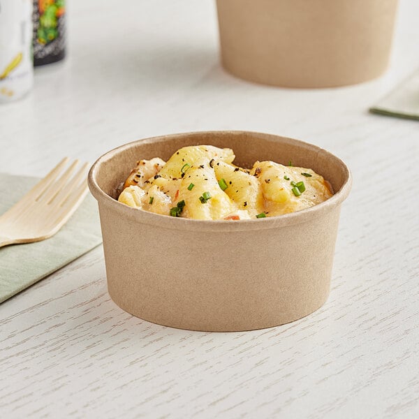 A bowl of macaroni and cheese in a Kraft paper food cup with a wooden fork on a table.