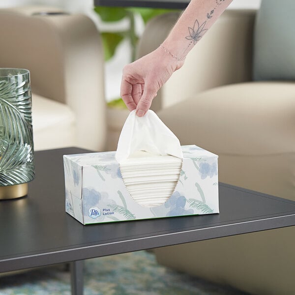 A hand holding a Puffs Plus Lotion facial tissue box on a table.