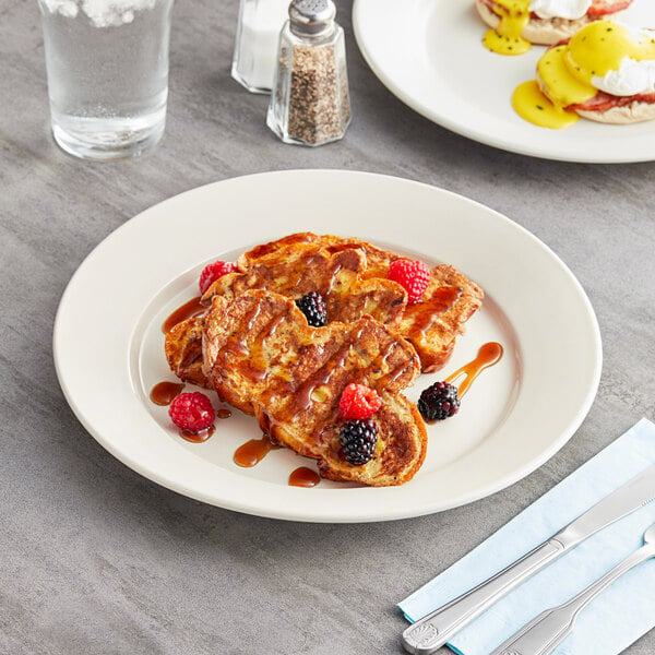 An Acopa ivory stoneware plate with French toast, berries, and syrup on a table with a glass of water.