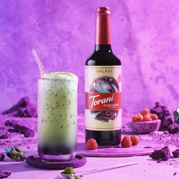 A close-up of a Torani Puremade Galaxy flavoring syrup bottle next to a glass of liquid.