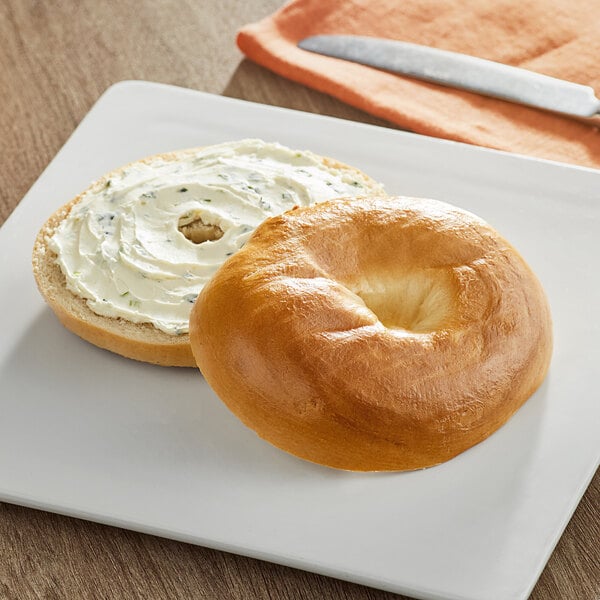 A Just Bagels plain bagel with cream cheese on a plate with a knife.