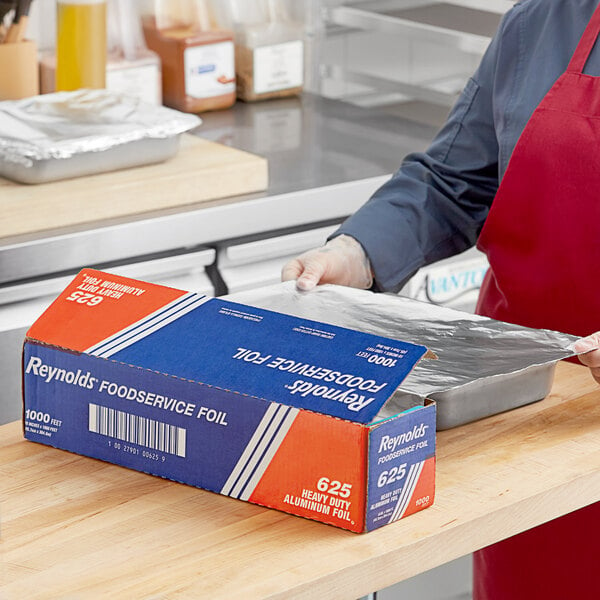 A person in a kitchen wearing a red apron and gloves cuts open a blue and white box of Reynolds Foodservice Heavy-Duty Aluminum Foil.